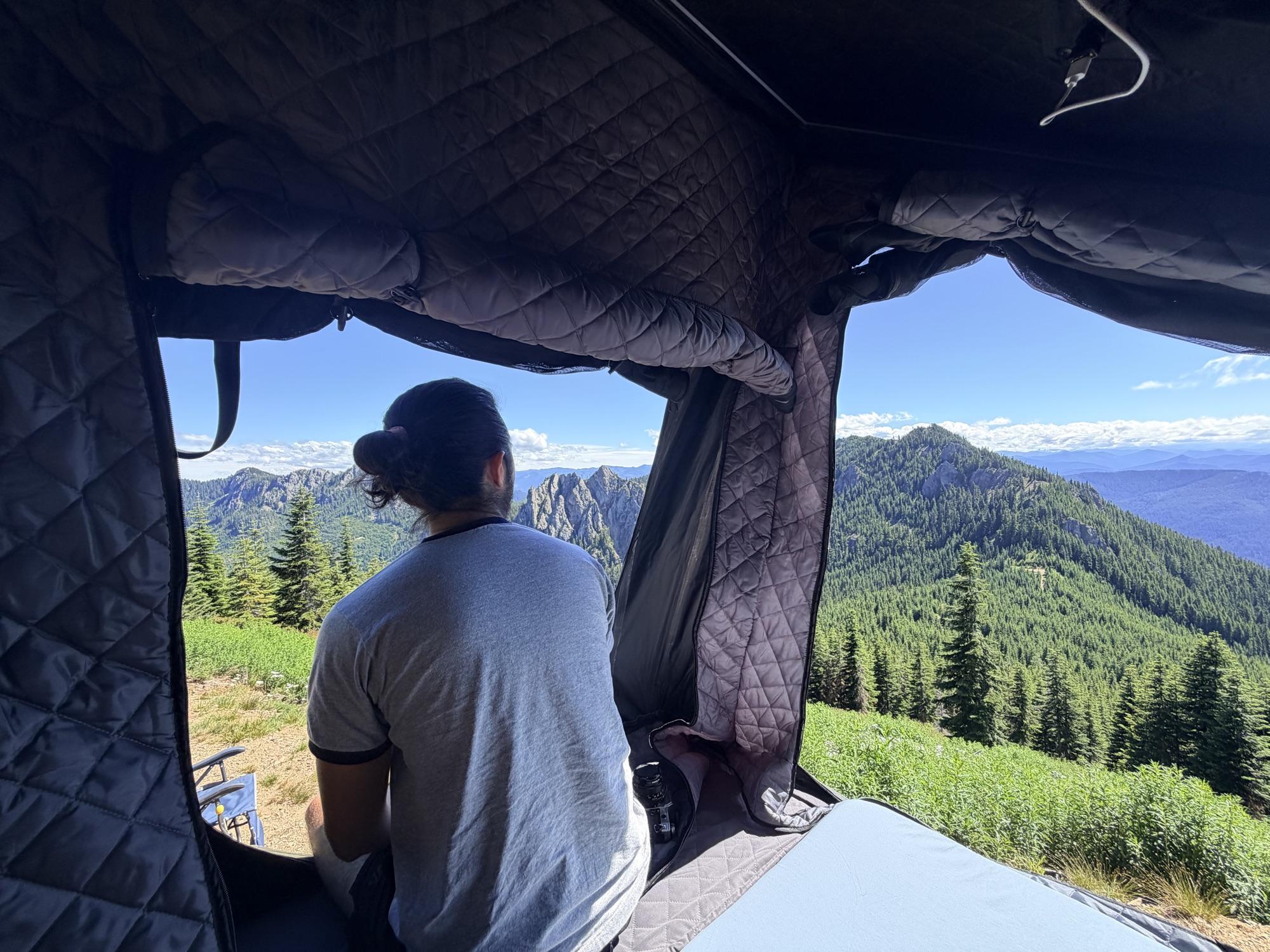 A camper sitting in the open doorway of a rooftop tent, looking out over forested mountains and rocky peaks