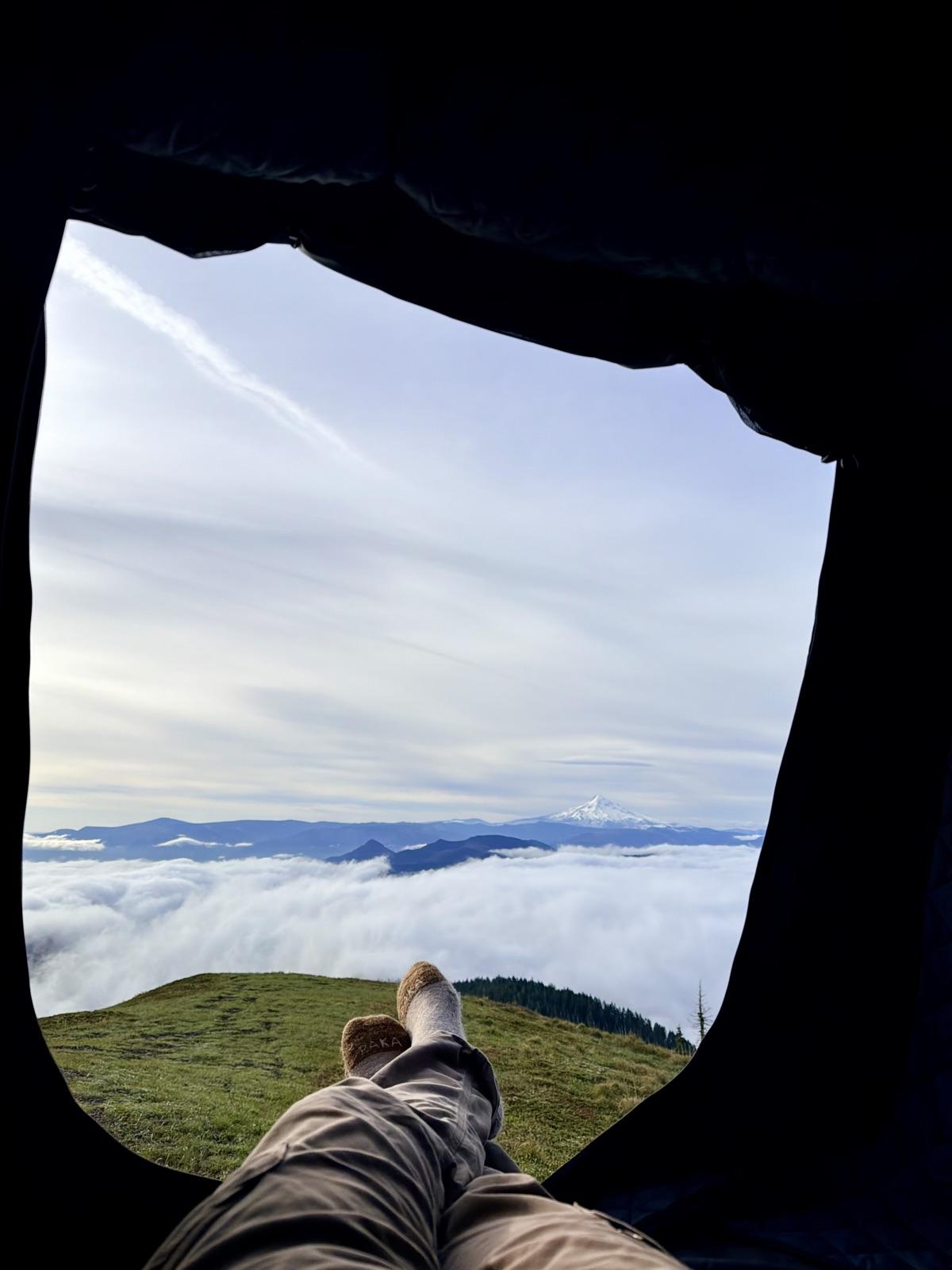 Socked feet resting in the open doorway of a rooftop tent, looking out over a sea of clouds to a snowcapped volcano