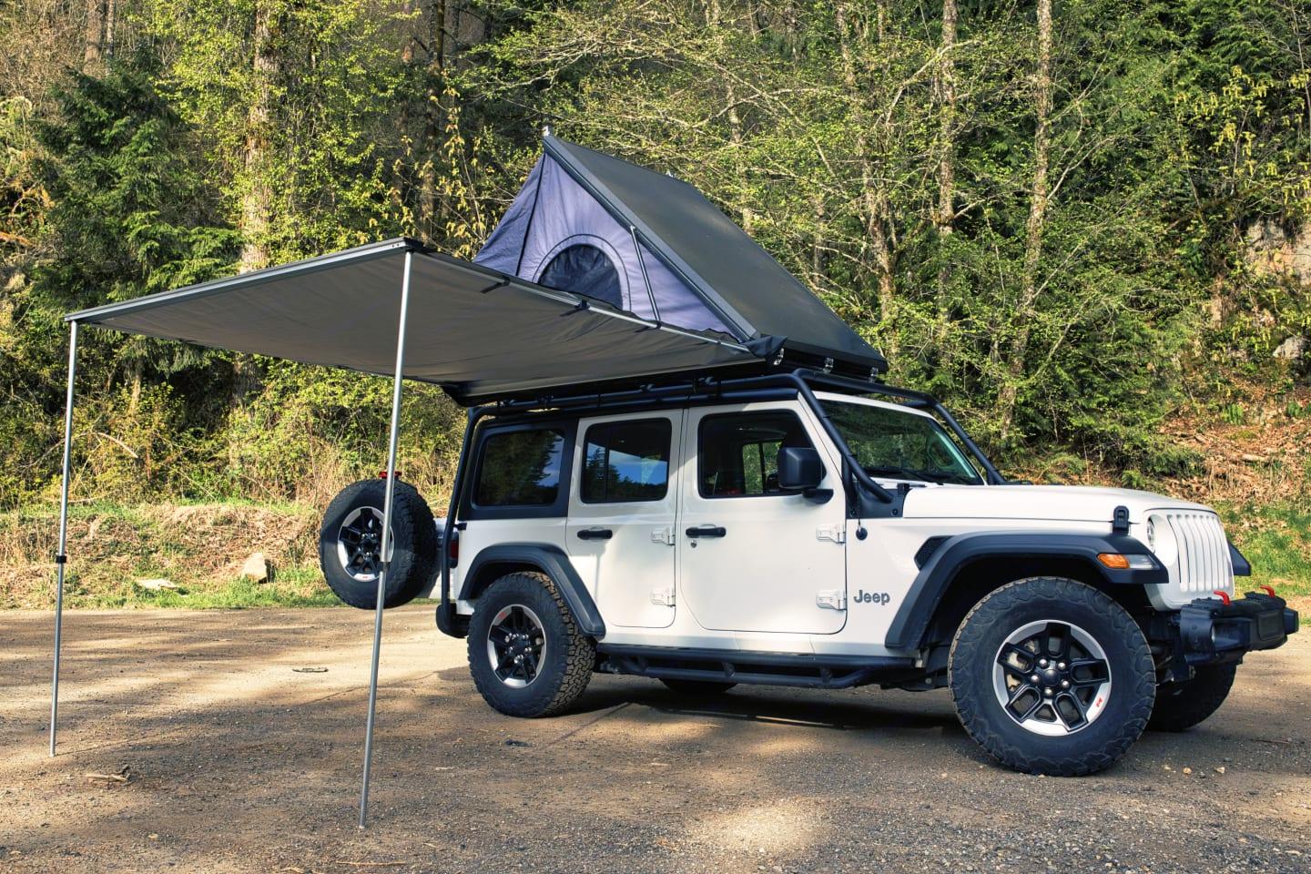 Jeep Wrangler with its rooftop tent open and side awning extended at a forest pull-out